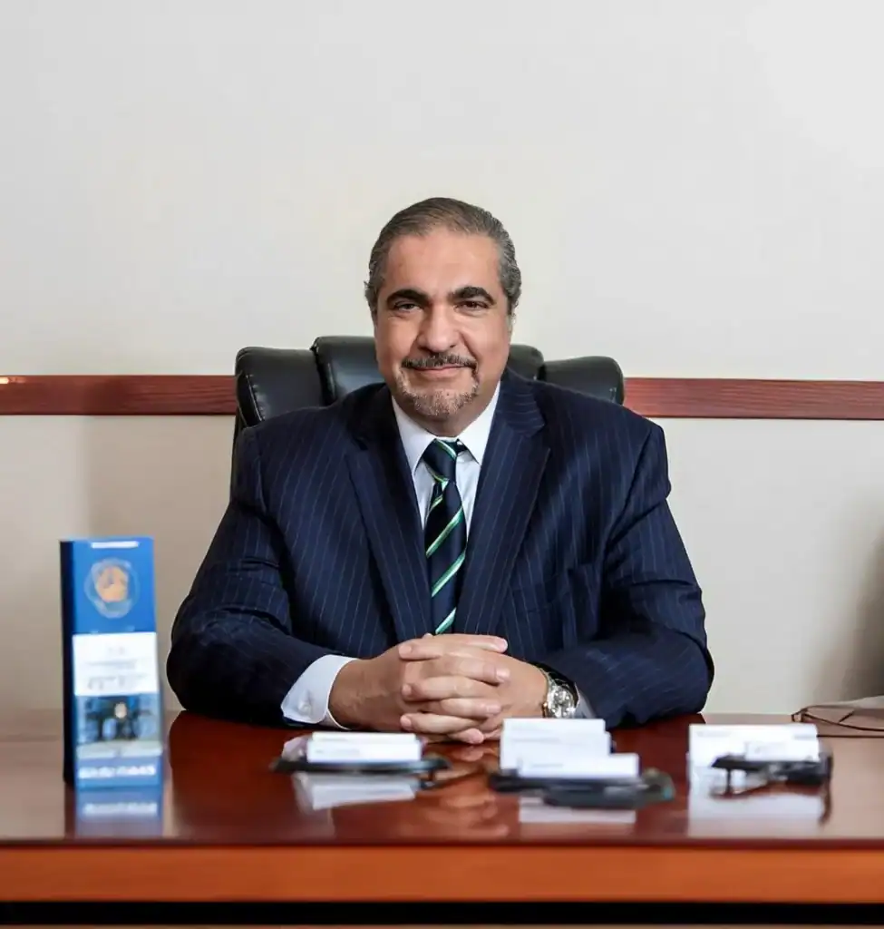 A Tony Rahnama in a pinstripe suit sits at a polished wooden desk, flanked by business cards and promotional materials.