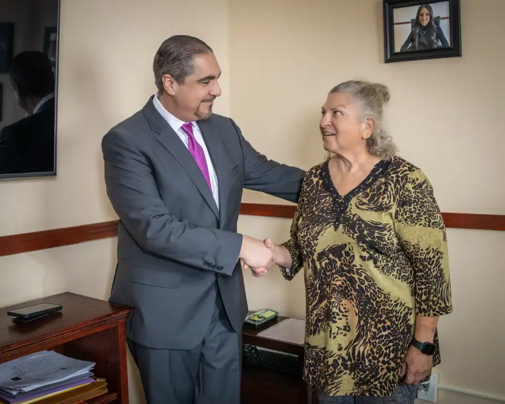 A man in a suit and a woman in a patterned shirt shake hands and smile at each other in an office setting.