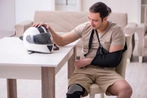 A man with an arm in a sling and a knee brace sits at a table, resting his hand on a motorcycle helmet and looking at it thoughtfully.