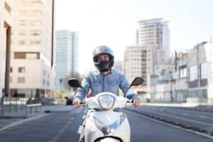 A person wearing a helmet and denim shirt rides a white scooter on an empty city street with tall buildings in the background.