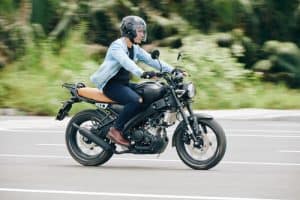 A person wearing a helmet and denim jacket rides a black motorcycle on a paved road with greenery in the background.