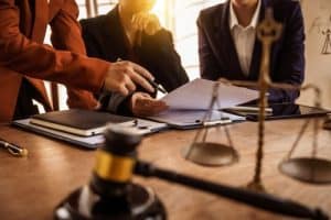 Three people in business attire review documents at a desk with a gavel, scales of justice, and notebooks.