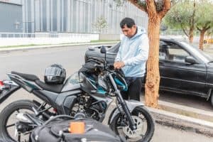 A man in a light blue hoodie stands by a parked motorcycle, adjusting a bag on the bike. A helmet rests on the motorcycle seat, and a black car is parked nearby.