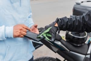 Person in a hoodie hands a wallet or document to a gloved individual standing beside a motorcycle on a street.