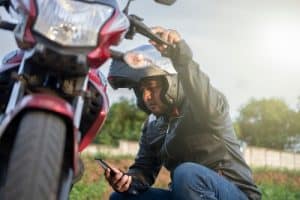 A man wearing a motorcycle helmet and leather jacket kneels beside a red motorcycle while looking at his phone outdoors.