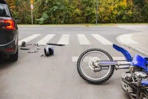 A motorcycle lies on its side near a crosswalk with scattered gear and a car parked nearby, suggesting a recent road accident.