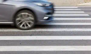 A gray car drives quickly through a pedestrian crosswalk on a city street, with the vehicle appearing slightly blurred from motion.