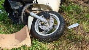 A damaged scooter lies on its side on grass, with the front wheel and fender visibly bent and broken.