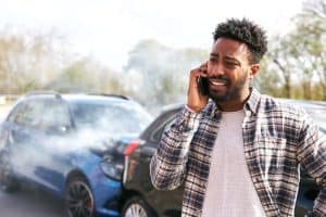 A man stands by two damaged cars after an accident, talking on his phone with smoke rising from the vehicles in the background.