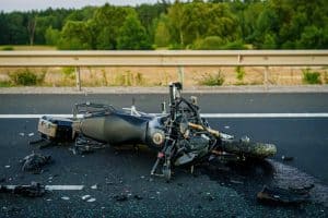 A damaged motorcycle lies on its side in the middle of a highway, surrounded by debris, with a metal guardrail and trees in the background.