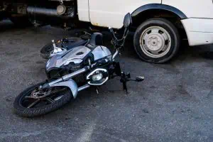 A silver motorcycle lies on its side on the pavement next to a white van with a damaged rear wheel.