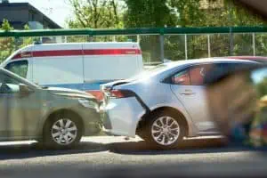 Two cars involved in a rear-end collision on a street, with an ambulance parked nearby and visible damage to both vehicles.