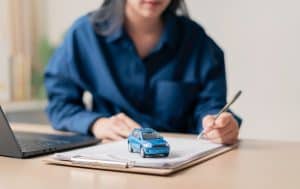 A person in a blue shirt writes on documents at a desk with a small blue toy car, a clipboard, and a laptop.
