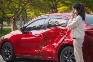 A woman stands next to a red car with a dented side, making a phone call, likely reporting an accident, in a sunny outdoor setting.