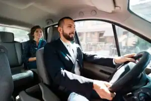 A man is driving a car while a woman sits in the back seat, both wearing seatbelts. Daylight enters through the windows.