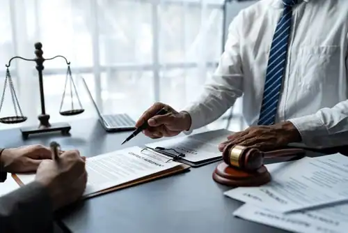 Two people in business attire review and sign legal documents at a desk with a gavel, scales of justice, and paperwork.