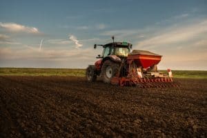 A red tractor with a seeder attachment is working on a plowed field under a partly cloudy sky.