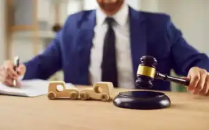 A person in a suit uses a gavel beside two wooden toy cars, suggesting a legal or judicial context related to automobiles.