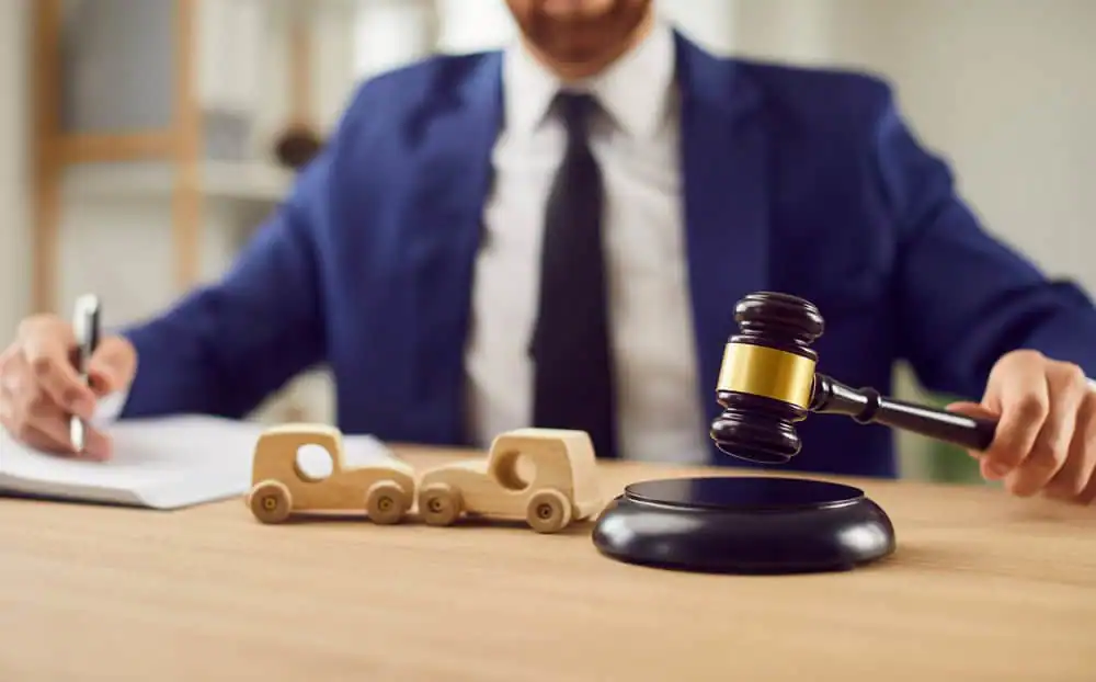 A person in a suit uses a gavel beside two wooden toy cars, suggesting a legal or judicial context related to automobiles.