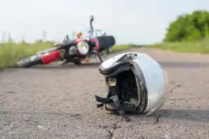 A motorcycle lies on its side in the background of a rural road, while a white helmet rests on the pavement in the foreground.