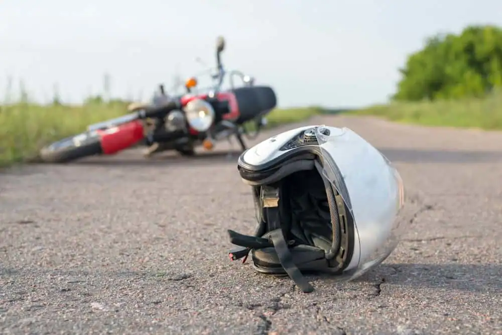 A motorcycle lies on its side in the background of a rural road, while a white helmet rests on the pavement in the foreground.
