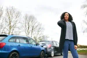 A woman stands beside two cars involved in a rear-end collision, holding her neck and appearing concerned.