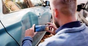 A man takes a photo of a large dent on the side of a blue car with his smartphone.