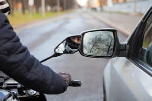 A motorcyclist wearing gloves and a jacket rides next to a car on a city street, with both vehicles' side mirrors visible in the foreground.