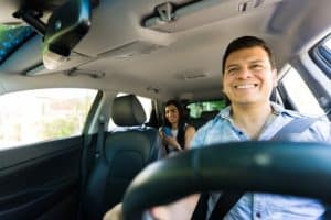 A man is driving a car, smiling, while a woman sits in the back seat looking at her phone.
