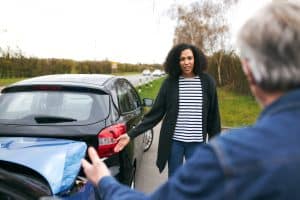 A woman points to the damage on her car’s rear bumper while talking to another person after a collision on the roadside.
