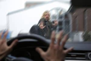 A woman with a shocked expression stands in front of a car with hands raised as the driver’s hands grip the steering wheel, viewed through the windshield.