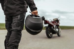 A person in black motorcycle gear holds a helmet while standing near a red motorcycle parked on an empty road.