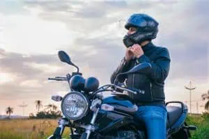 A person wearing a black helmet and jacket sits on a motorcycle outdoors, fastening their helmet strap with one hand.