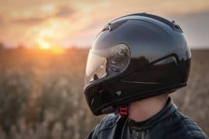 Person wearing a black motorcycle helmet and leather jacket standing outdoors, with a blurred field and sunset in the background.