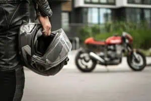 Person in black leather jacket holding a motorcycle helmet, with a red motorcycle parked on the street in the background.