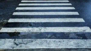 A crosswalk with white stripes on wet asphalt; the lower stripes show signs of wear and cracks in the pavement.