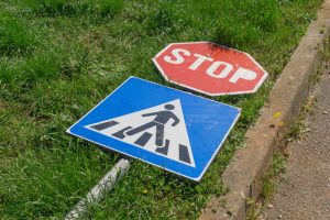 A stop sign and a pedestrian crossing sign lie on the grass next to a curb, both knocked over and not standing upright.