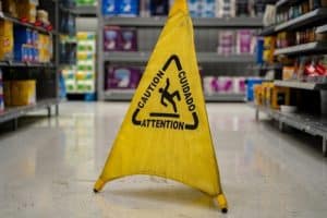A yellow caution sign warning of a wet floor stands in the aisle of a store with shelves of products in the background.