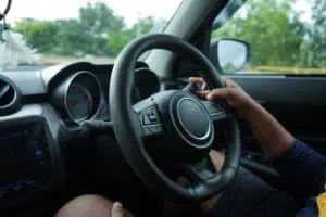 Person's hands holding the steering wheel of a car while driving, interior view with dashboard and window showing greenery outside.