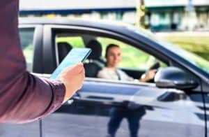 Person holding a smartphone on the street approaches a parked car with a smiling driver inside.