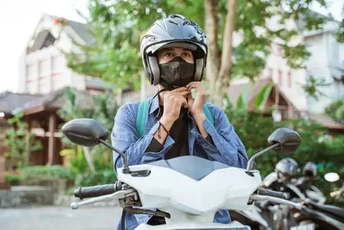 A person wearing a helmet and face mask fastens the strap while sitting on a white motorbike outdoors.