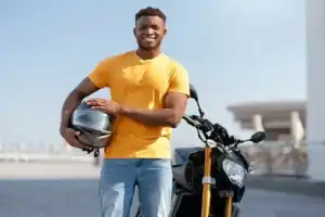 A man in a yellow t-shirt stands outdoors holding a motorcycle helmet next to a black motorcycle, smiling at the camera.