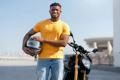 A man in a yellow t-shirt stands outdoors holding a motorcycle helmet next to a black motorcycle, smiling at the camera.