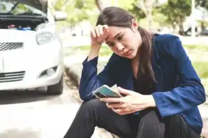 A woman sits on a curb looking worried, holding her phone and head, with a car parked nearby and its hood open.