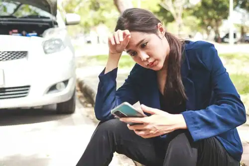 A woman sits on a curb looking worried, holding her phone and head, with a car parked nearby and its hood open.