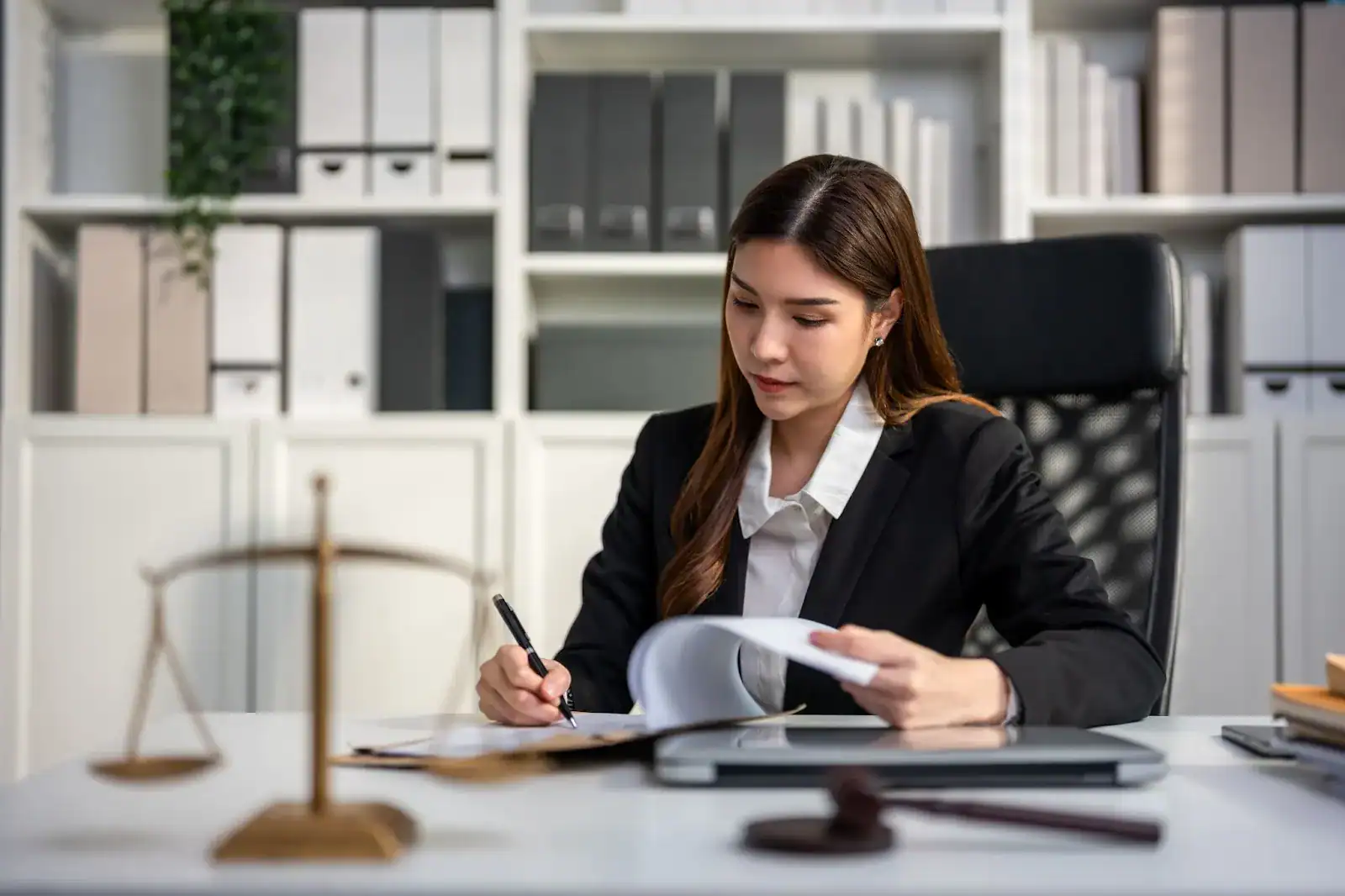 A woman in professional attire sits at a desk reviewing documents, with legal scales and a gavel on the table, in an office with shelves of files and books.
