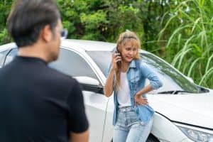 A woman stands by a damaged white car, talking on her phone, while a man in a black shirt stands in the foreground.