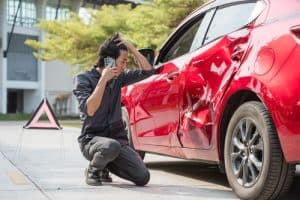 A person kneels beside a red car with a damaged side, talking on a phone. A warning triangle is placed behind the vehicle.
