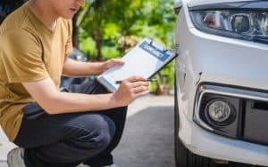 A person holding a clipboard labeled "Contract" inspects and writes notes near a dent on the front bumper of a white car.