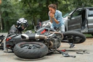 A motorcyclist sits on the ground next to a fallen motorcycle after a crash, while a concerned woman stands nearby and a car with an open door is visible in the background.